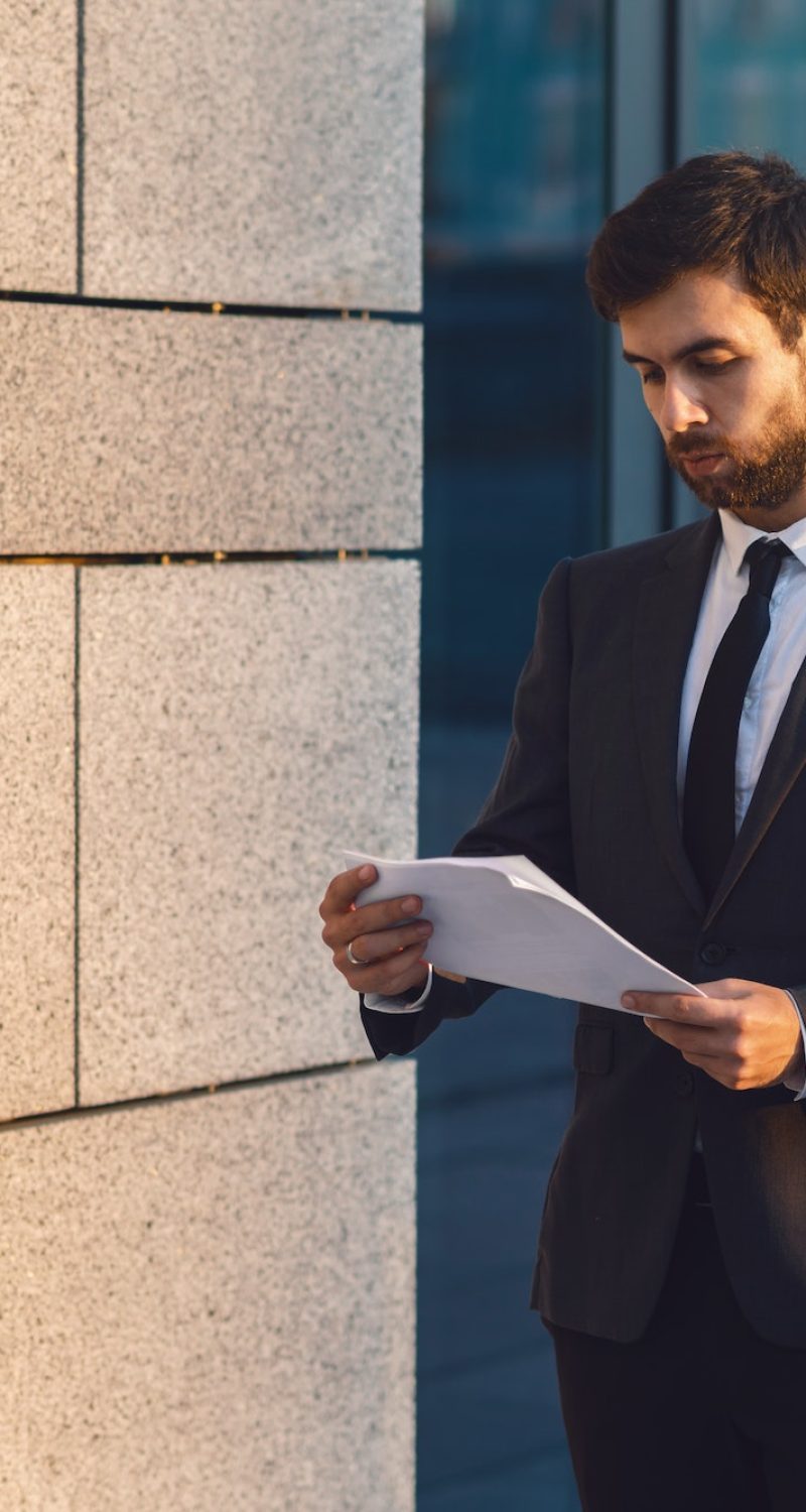 Serious young businessman in a classic suit reading advantageous business document in paper